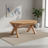 A light natural wood oval dining table with a geometric X-shaped base, shown in a modern room setting on a textured beige rug with a potted plant and wall art in the background.