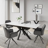 A modern dining room scene featuring a rectangular white marble-top dining table with a black metal X-frame base, surrounded by four gray upholstered swivel chairs on a light gray rug. The room has light gray walls, minimalist framed wall art, and a side console table in the background.