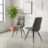 Two modern gray upholstered dining chairs with black metal legs, shown in a bright minimalist room with a potted plant and framed wall art.