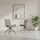 Two modern swivel dining chairs upholstered in light taupe fabric with black metal legs, arranged in a minimalist room setting with a framed abstract print on the wall and a potted plant to the right.