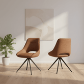 Two modern upholstered swivel chairs in warm brown tan faux leather with black metal tripod legs, shown in a bright minimalist room with a light wood floor, a potted plant, and abstract wall art.