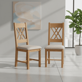 Two light oak dining chairs with cream upholstered seats and crossed backrests, shown in a bright modern room with a plant and abstract wall art.