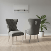 Two dark grey Velvet Fabric upholstered dining chairs with silver metal legs and nailhead trim, Lion Knocker back shown in a styled room with a potted plant and wall art.