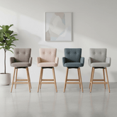 Four modern Cotswold Counter stools with tufted fabric upholstery in gray, blush pink, slate blue, and light gray, all with natural wood legs, arranged in a row against a minimalist white wall with a framed abstract print and a potted plant.