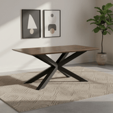 A rectangular dining table with a medium brown wood grain top and a black metal X-shaped base, styled on a textured beige rug in a modern room setting.