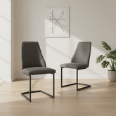Two modern gray upholstered dining chairs with black metal cantilever legs, shown in a bright minimalist room with light wood flooring and a potted plant.