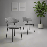 Two modern upholstered dining chairs with curved light gray fabric seats and backs and dark metal legs, shown in a minimalist gray room with two framed prints and a potted plant.