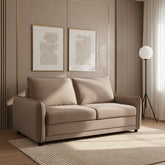A two-seater sofa bed upholstered in a taupe-beige microfiber fabric, styled in a bright, modern living room with a light rug, floor lamp, and white bookshelf.