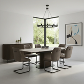 A modern dining room setup featuring a dark brown ribbed rectangular table with a light stone top, surrounded by six taupe upholstered chairs with black metal sled bases. The room has white walls, a large black-framed window looking out to green trees, a black multi-light chandelier, and a matching dark brown ribbed sideboard with small potted plants.