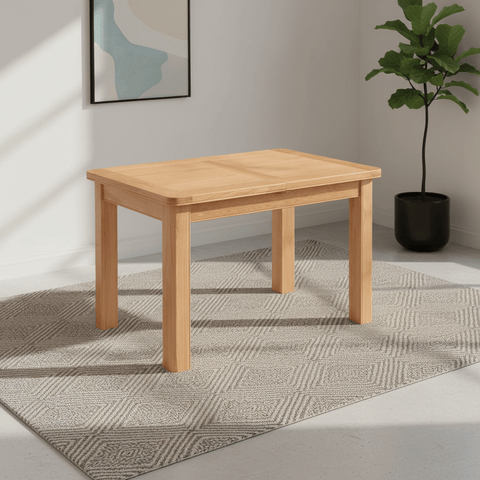 A light natural wood Chatsworth Oak rectangular table with four legs, shown in a styled room setting on a textured gray rug with a potted plant and framed wall art in the background.