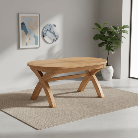 A light natural wood oval dining table with a geometric X-shaped base, shown in a modern room setting on a textured beige rug with a potted plant and wall art in the background.