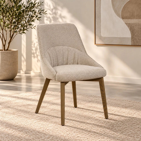 A modern upholstered dining chair with a textured light beige fabric seat and back, supported by tapered medium-brown wooden legs, shown in a styled room setting with a potted plant and abstract wall art.