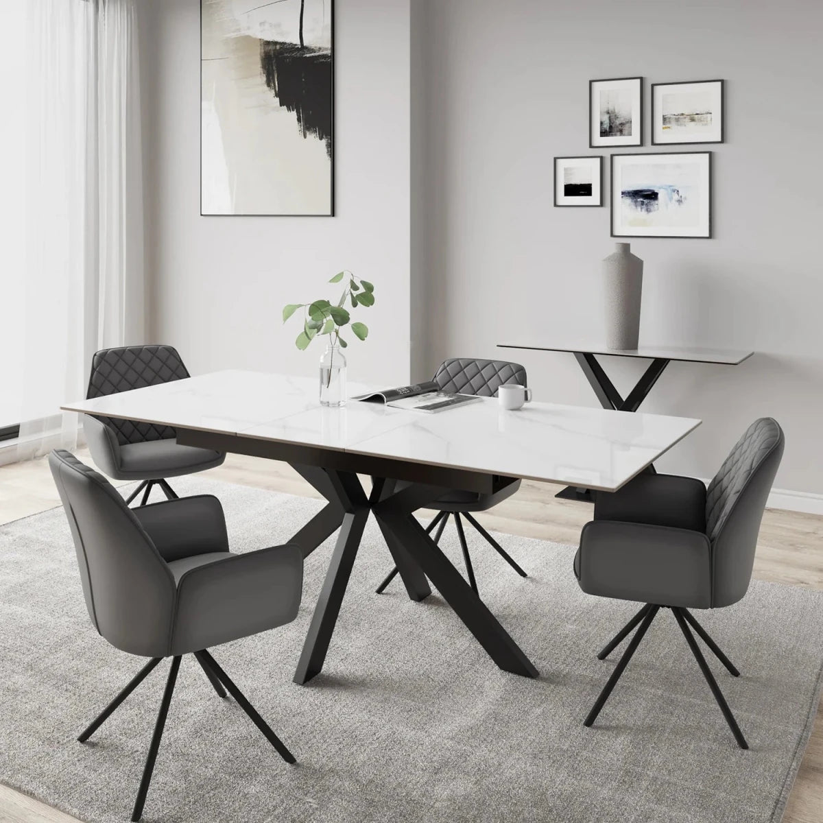 A modern dining room scene featuring a rectangular white marble-top dining table with a black metal X-frame base, surrounded by four gray upholstered swivel chairs on a light gray rug. The room has light gray walls, minimalist framed wall art, and a side console table in the background.