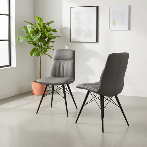 Two modern gray upholstered dining chairs with black metal legs, shown in a bright minimalist room with a potted plant and framed wall art.