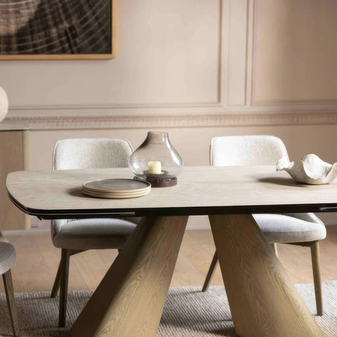 A modern dining table with a cream ceramic top and sculptural angled wooden legs, surrounded by upholstered dining chairs in a softly lit dining room.