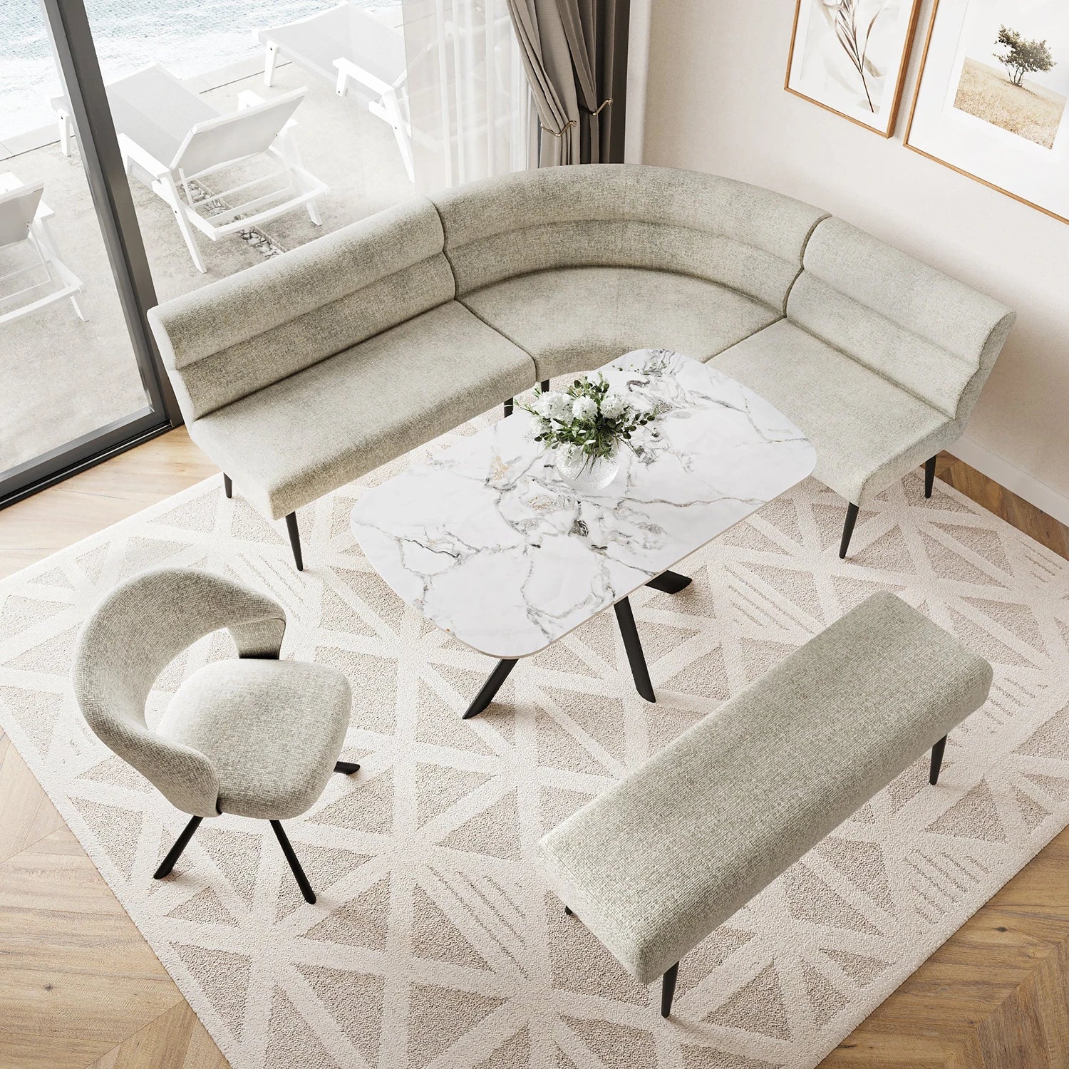 A modern curved U-shaped upholstered sofa in a light beige fabric, paired with a matching rectangular bench, a curved accent chair, and a white marble-top coffee table on a geometric patterned rug in a bright living room.