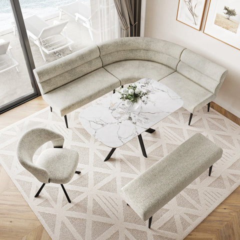A modern curved U-shaped upholstered sofa in a light beige fabric, paired with a matching rectangular bench, a curved accent chair, and a white marble-top coffee table on a geometric patterned rug in a bright living room.