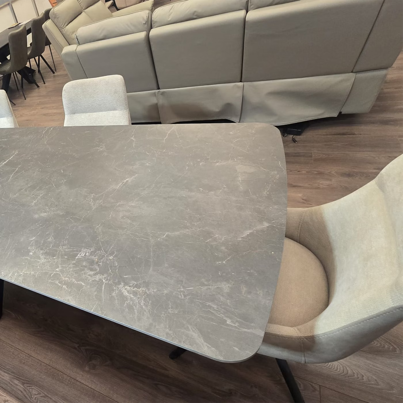 Beatrix Slate Mist Sintered Stone Extending Dining Table with a curved front edge, paired with light beige upholstered chairs in a furniture showroom with a gray leather sofa in the background.