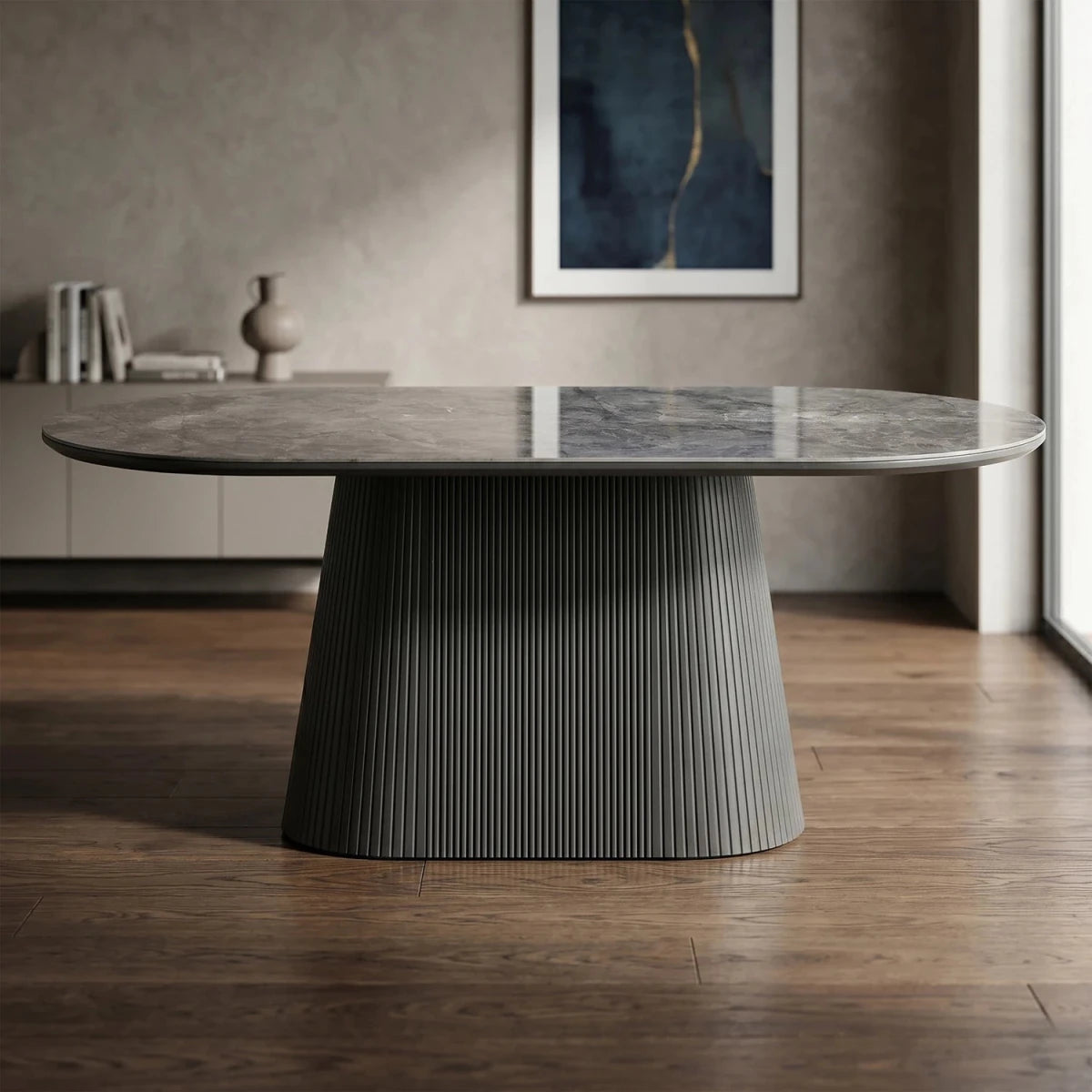A modern oval dining table with a polished gray stone top and a sculptural fluted pedestal base, set in a contemporary living room with wood floors and a textured wall.