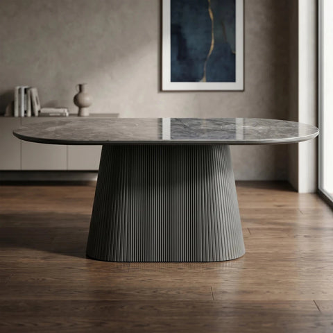 A modern oval dining table with a polished gray stone top and a sculptural fluted pedestal base, set in a contemporary living room with wood floors and a textured wall.