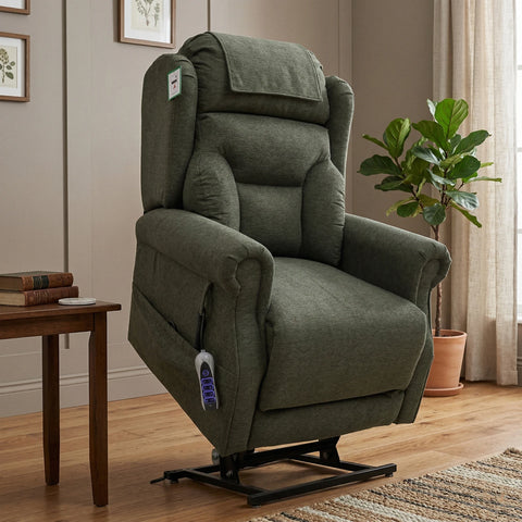 An upholstered recliner chair in a muted sage green fabric, shown in a staged living room setting with a side table, framed wall art, a potted plant, and a window curtain.