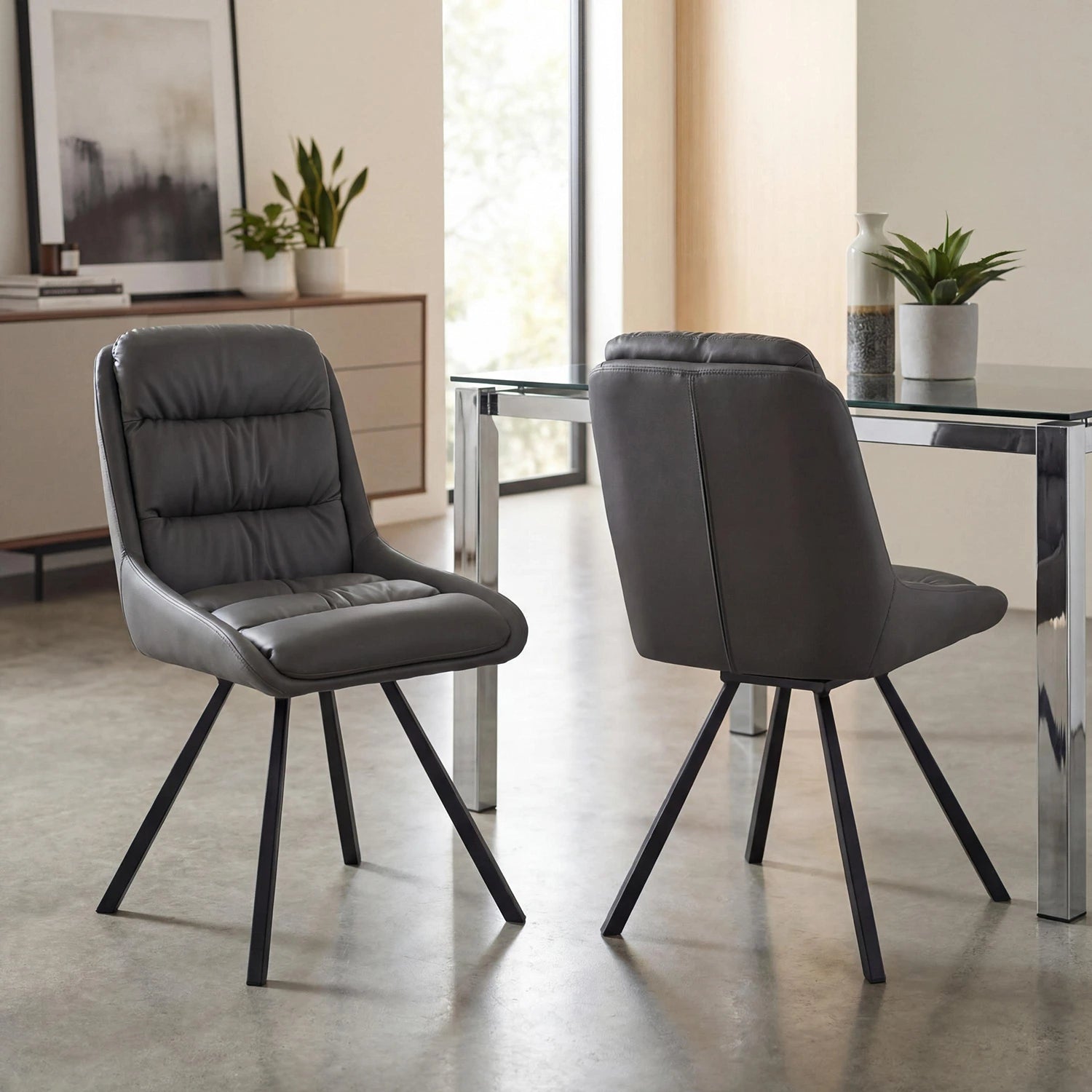 Two modern gray upholstered dining chairs with black metal legs, shown in a bright contemporary dining room next to a glass-top table.