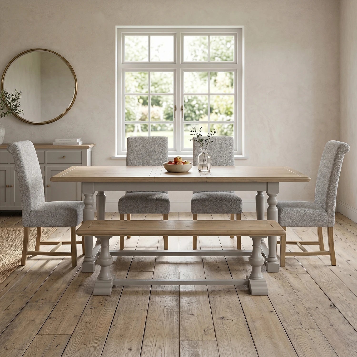 A light oak dining table set with four light gray upholstered chairs, a matching wooden bench, and a small vase of flowers on a rustic wood floor in a bright dining room.