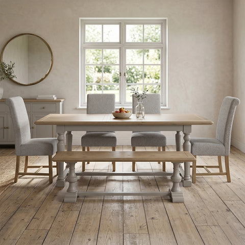 A light oak dining table set with four light gray upholstered chairs, a matching wooden bench, and a small vase of flowers on a rustic wood floor in a bright dining room.