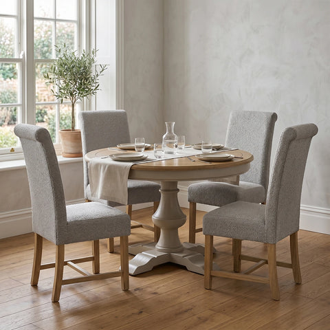 A round wooden dining table with a pedestal base, surrounded by four upholstered dining chairs in a bright room with a window, wood floor, and potted plant.