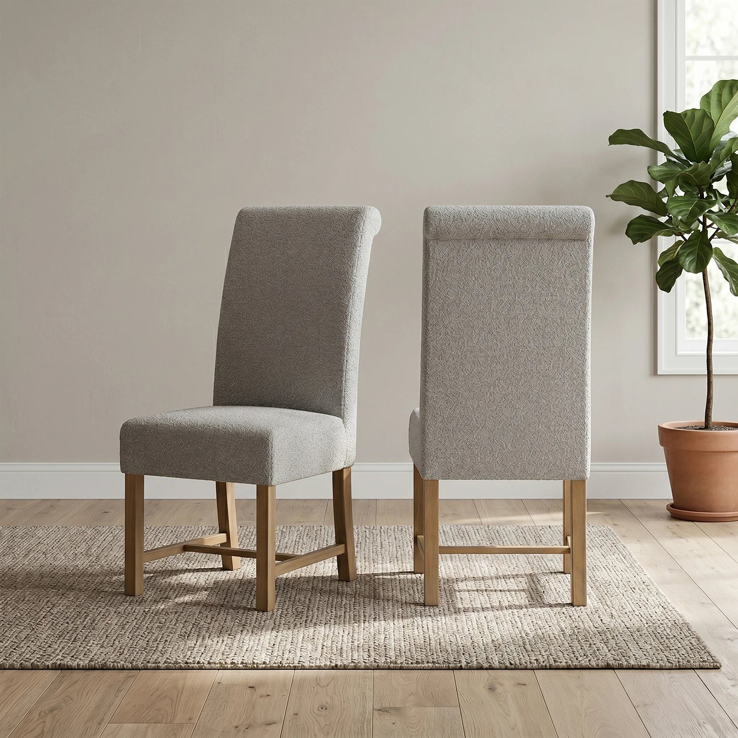 Two upholstered dining chairs with light gray fabric and natural wood legs, shown from the front and back on a woven rug in a bright room with a potted plant.
