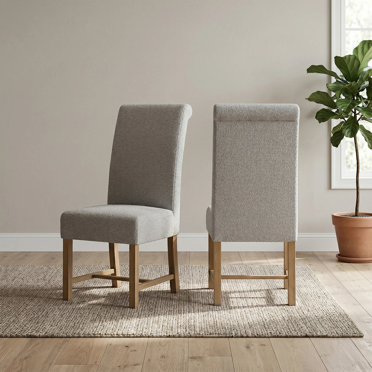 Two upholstered dining chairs with light gray fabric and natural wood legs, shown from the front and back on a woven rug in a bright room with a potted plant.
