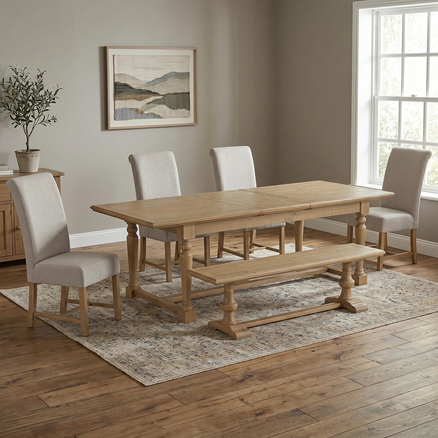 A light oak wood dining table set in a dining room with four light beige upholstered chairs, a matching wooden bench, and a patterned area rug.