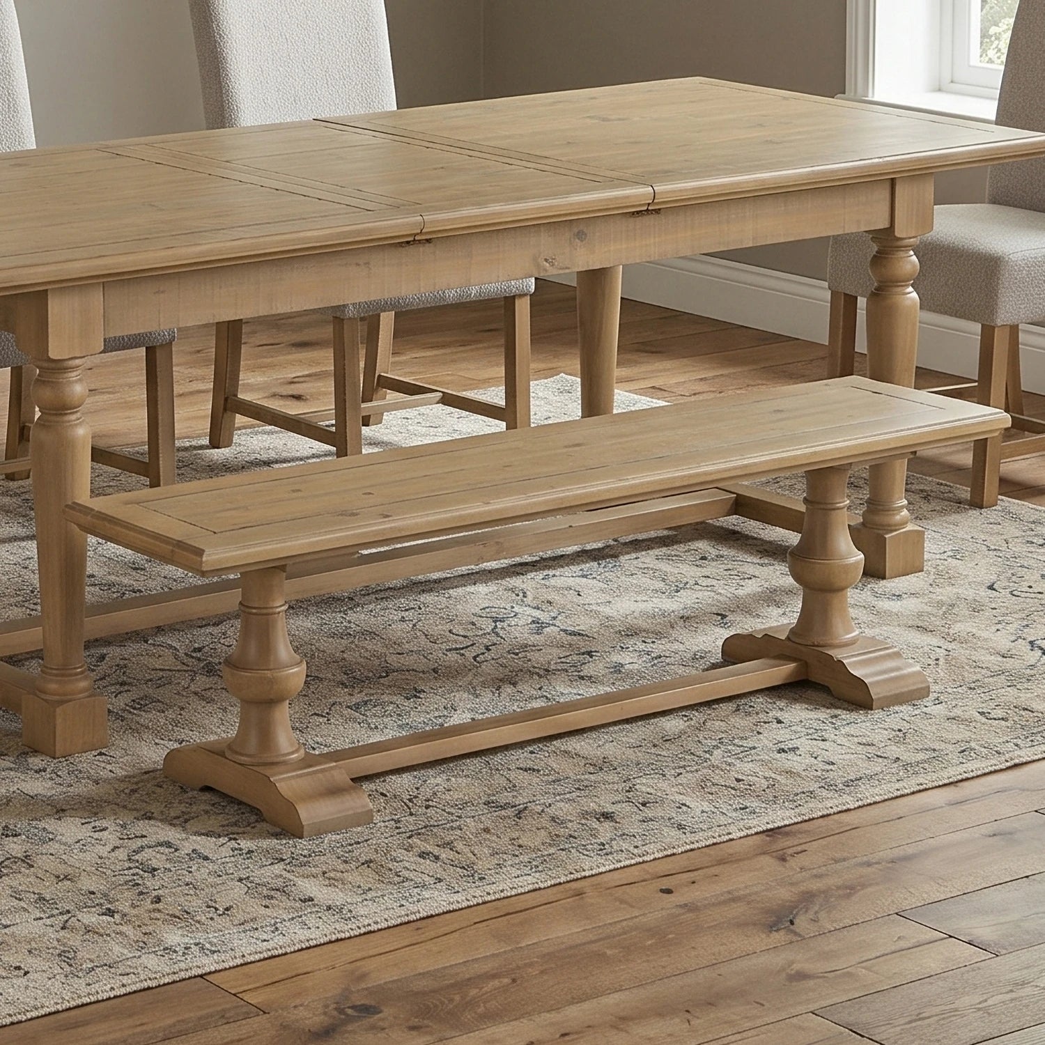 A light natural oak dining table with matching wooden bench, set on a patterned area rug in a dining room with neutral chairs.