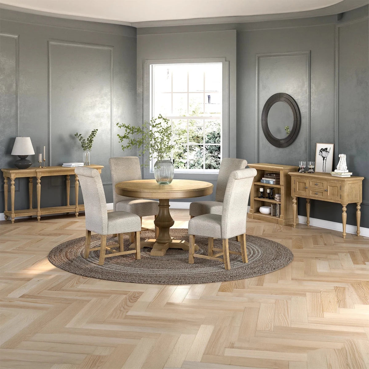 A round wooden dining table with four upholstered cream chairs sits on a round textured rug in a styled dining room. The room features gray paneled walls, a light wood herringbone floor, and matching wooden sideboards and a console table.