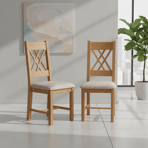 Two light oak dining chairs with cream upholstered seats and crossed backrests, shown in a bright modern room with a plant and abstract wall art.