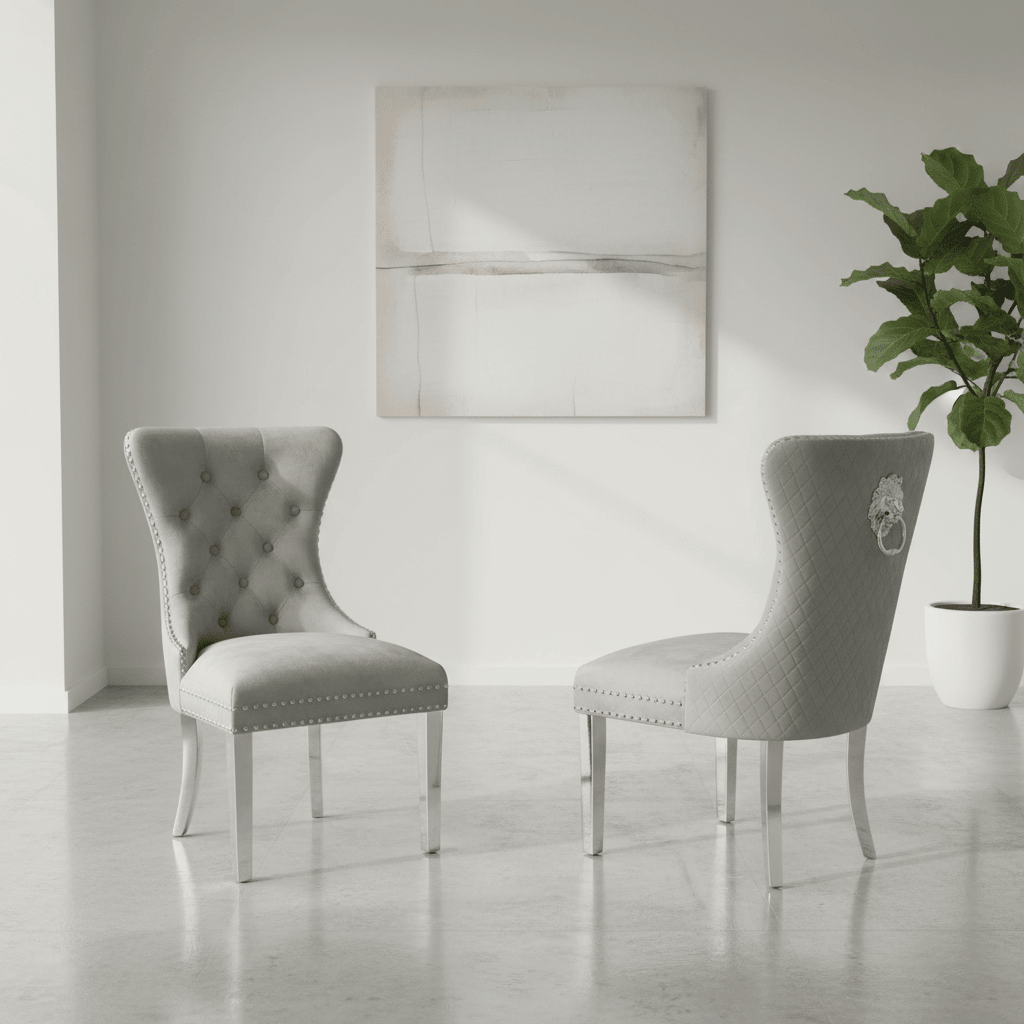 Two light gray upholstered dining chairs with silver nailhead trim and white legs, shown in a bright minimalist room with a potted plant and abstract wall art.