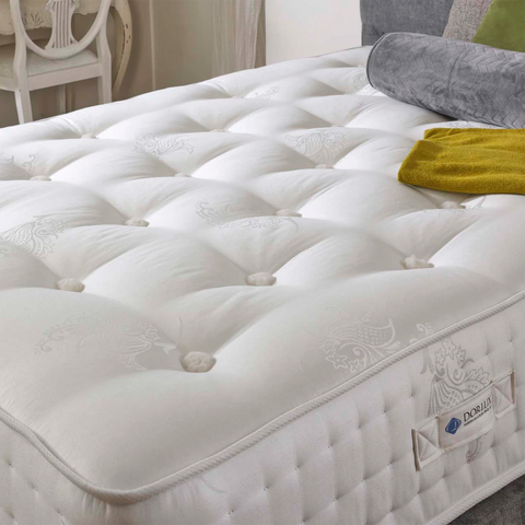 A close-up of a white tufted mattress with decorative button tufting and a visible DORLUX brand label on the side, styled in a bedroom setting with pillows and a folded blanket.
