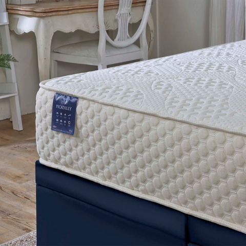 A close-up of a mattress corner with a textured cream quilted top, a blue 'PICADILLY' By Dorulx brand tag, and a dark navy bed base in a softly styled bedroom setting.