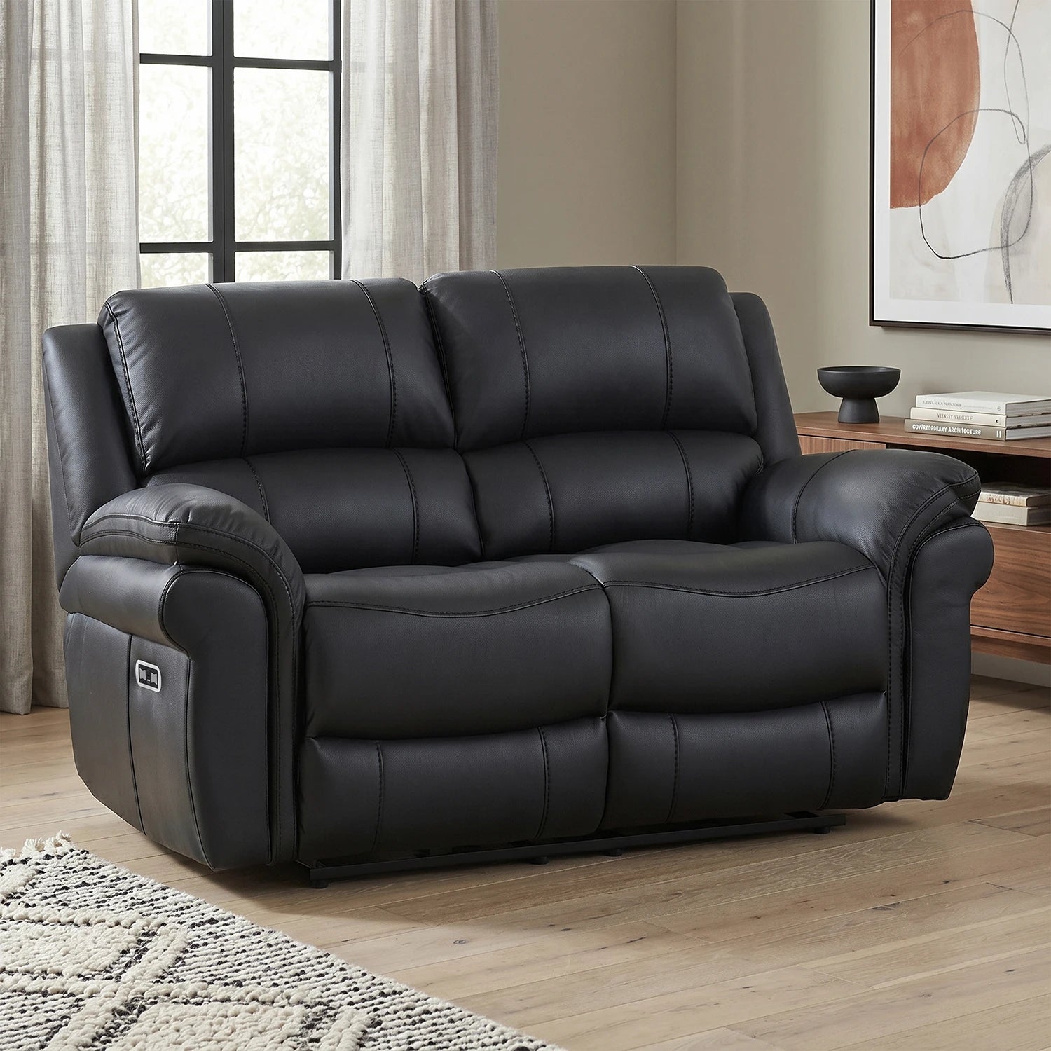 Black leather reclining 2 Seater sofa in a modern living room setting with wood floors, a sideboard, and a window in the background.