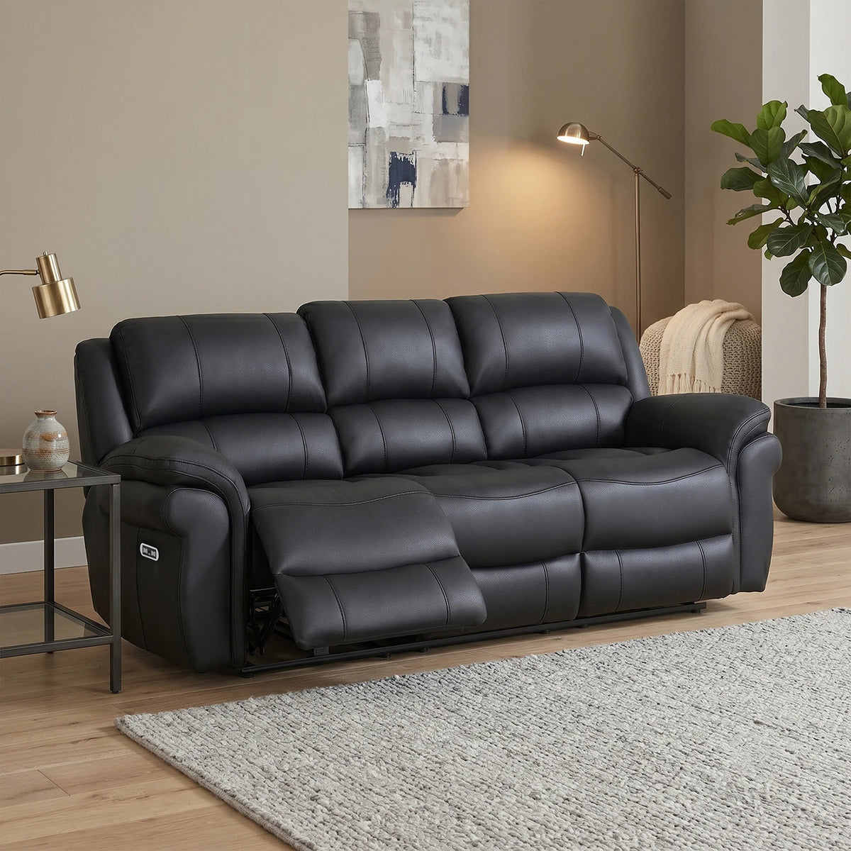 A black leather reclining sofa shown in a styled living room setting with a side table, floor lamp, wall art, and a potted plant.