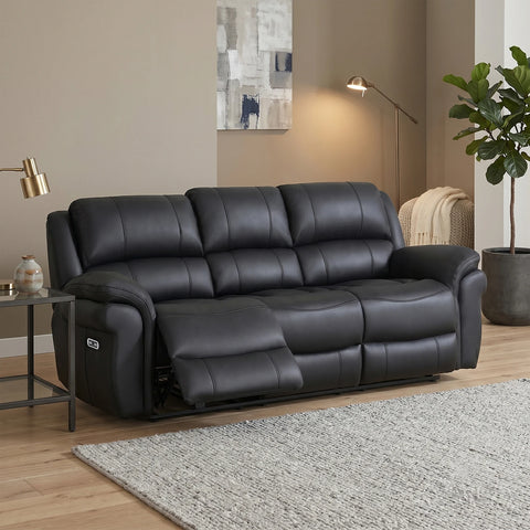A black leather reclining sofa shown in a styled living room setting with a side table, floor lamp, wall art, and a potted plant.