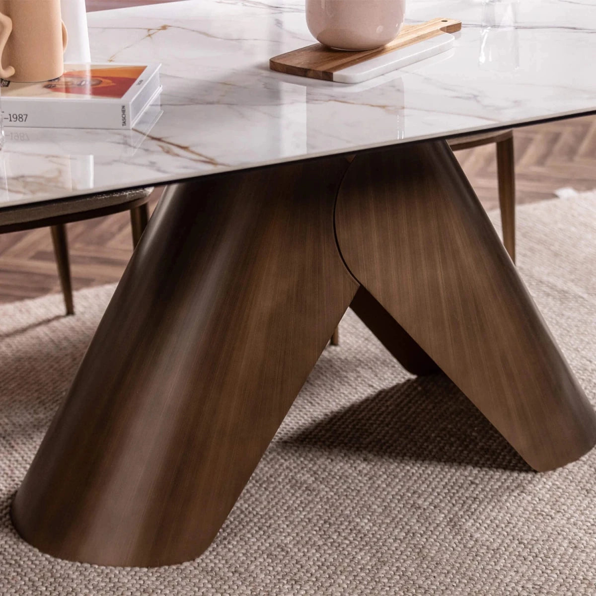A close-up of a modern dining table featuring a glossy dark brown Metal A-frame base and a white marble-patterned ceramic tabletop, styled on a textured beige rug with books and a vase in the background.