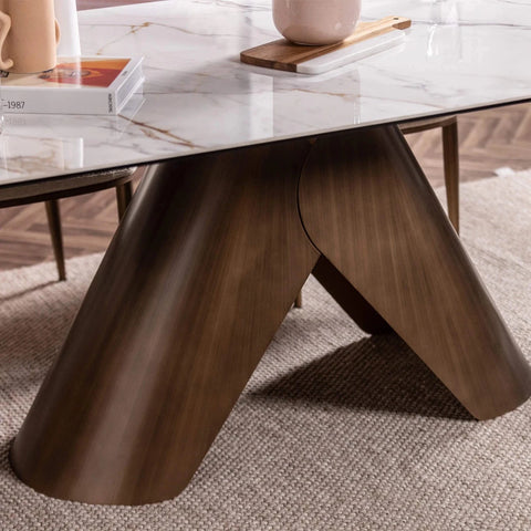 A close-up of a modern dining table featuring a glossy dark brown Metal A-frame base and a white marble-patterned ceramic tabletop, styled on a textured beige rug with books and a vase in the background.