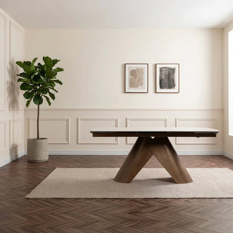 A modern dining table with a white top and angled wooden legs is styled in a minimalist dining room setting with a beige rug, wooden herringbone floor, and a pair of large abstract wall art panels behind it.