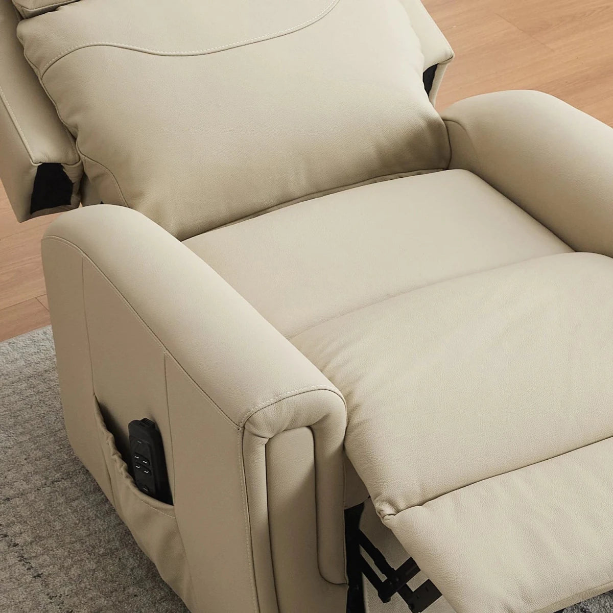 A beige leather recliner chair shown in a close-up angle, with the seat extended and a black remote control in the side pocket, set on a gray rug over a light wood floor.