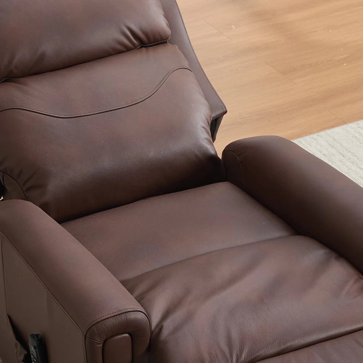 A close-up of a brown leather recliner chair showing the padded backrest, armrests, and seat cushion, set against a light wood floor and a small rug.