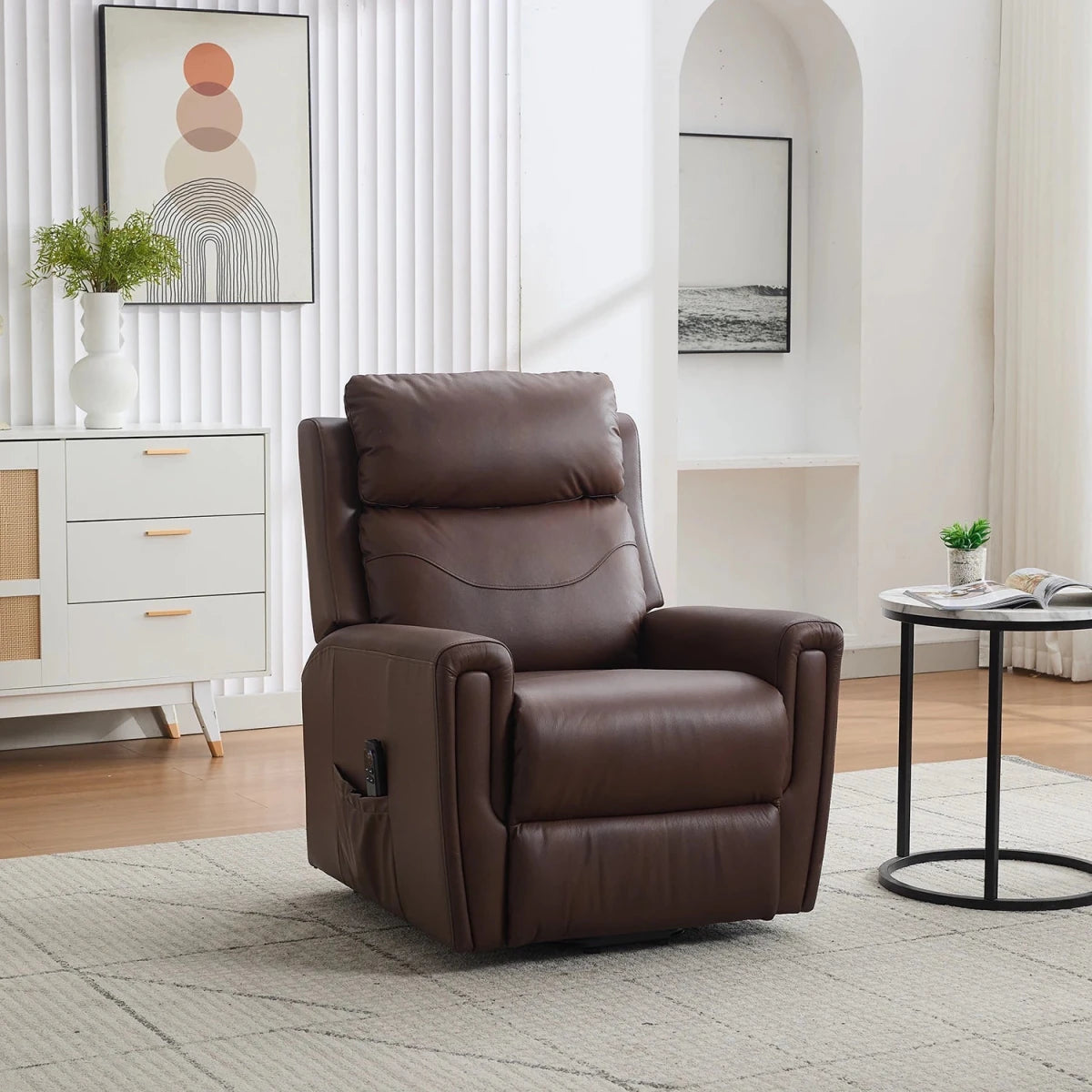 A dark brown leather riser recliner chair with a headrest and side pocket, set in a bright, modern living room next to a white sideboard and a small round table.