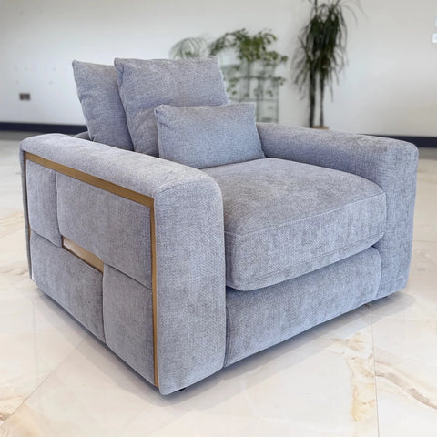 A modern gray upholstered armchair with a square base, wide armrests, and a slim gold-toned metal frame accent, shown in a minimalist indoor room setting.