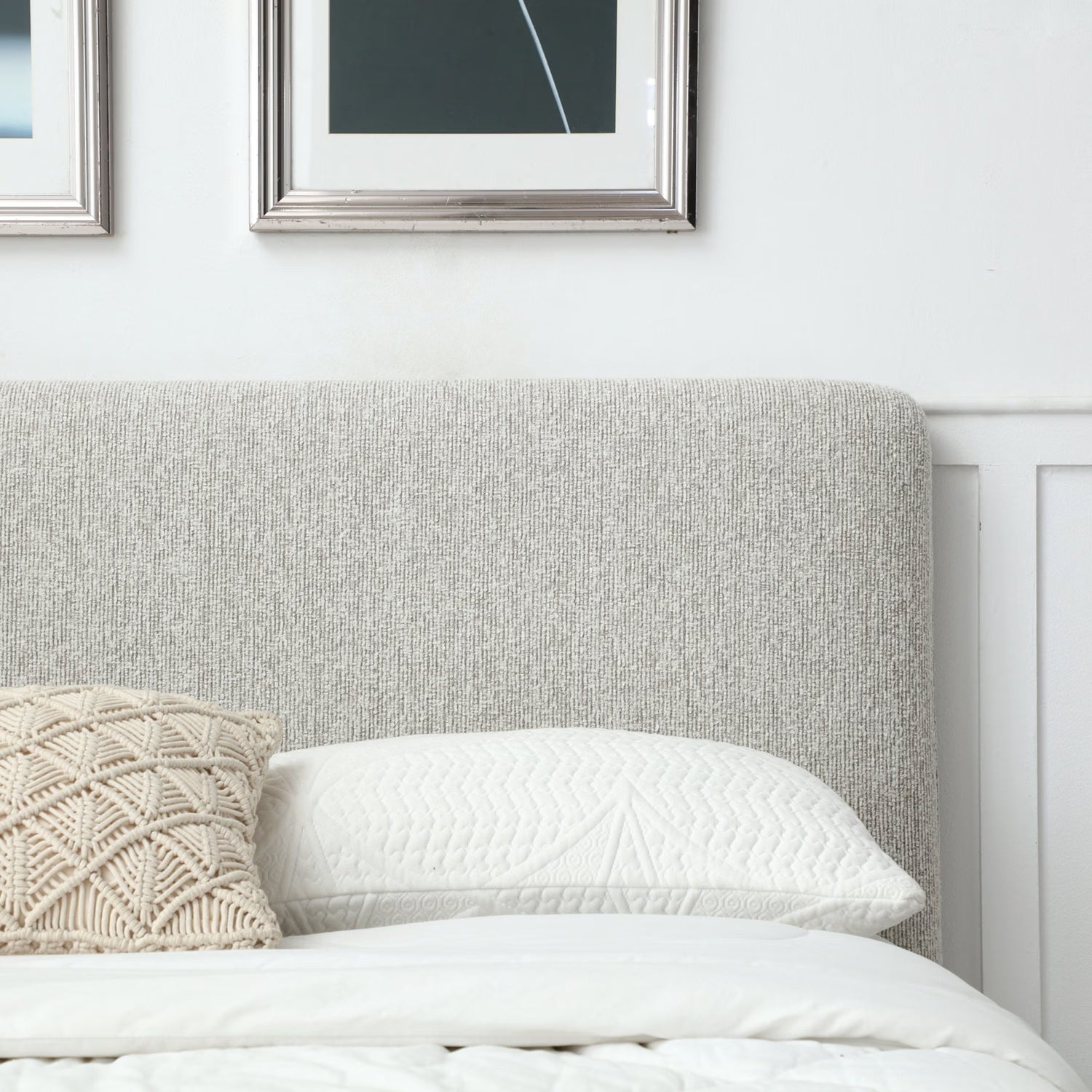 A close-up of a bed with a tall upholstered headboard in a light gray textured fabric, styled with white bedding, a white pillow, and a cream woven accent pillow. Framed wall art is partially visible above on a white wall.