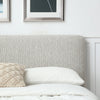 A close-up of a bed with a tall upholstered headboard in a light gray textured fabric, styled with white bedding, a white pillow, and a cream woven accent pillow. Framed wall art is partially visible above on a white wall.